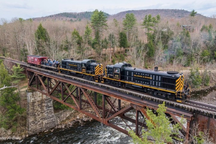 Black train crosses a scenic bridge over a river, surrounded by trees and hills.