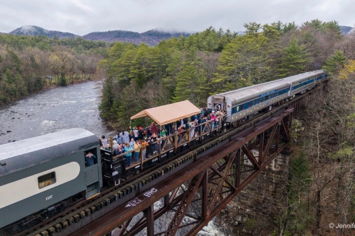 Train with open passenger car crosses a forested bridge over a river.