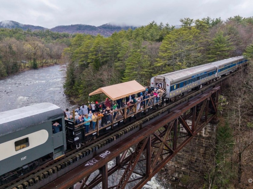 Train with open passenger car crosses a forested bridge over a river.