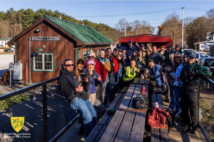 Group of people giving thumbs up outside a small building with a green roof.