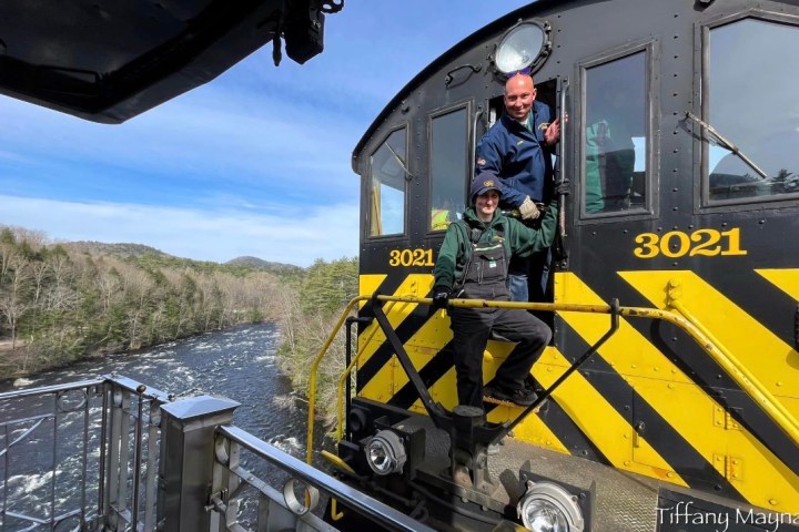 Two people in train cabin above river with trees in the background, engine number 3021 visible.