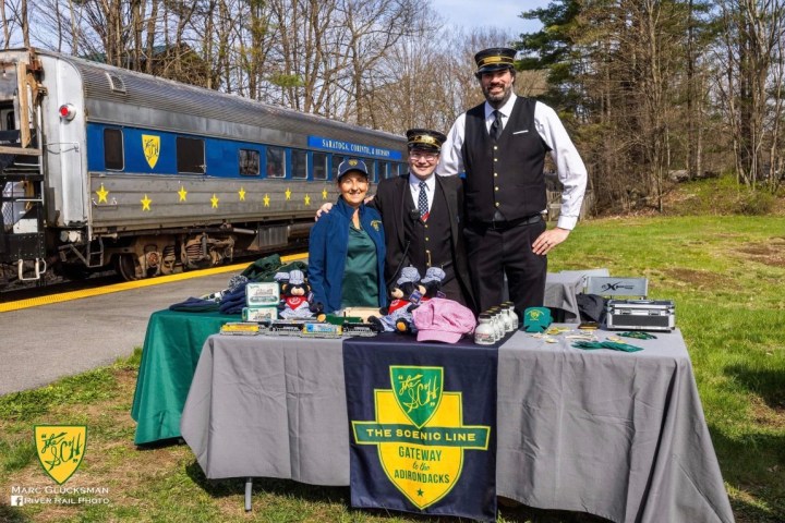 Three people in conductor uniforms stand by a table with train models and merchandise near a train.
