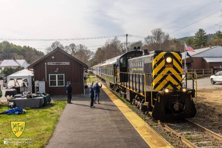 Black train at station with people waving, Hadley/Luzerne sign, and American flag visible.