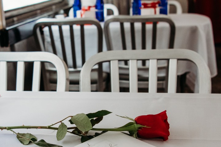Red rose and envelope on a table with chairs and bottles in the background.
