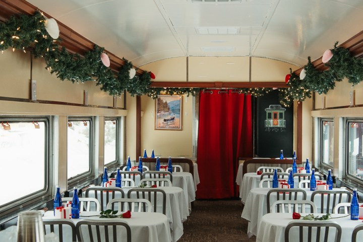 Train dining car with white tablecloths, red roses, and blue bottles on tables, decorated with greenery.