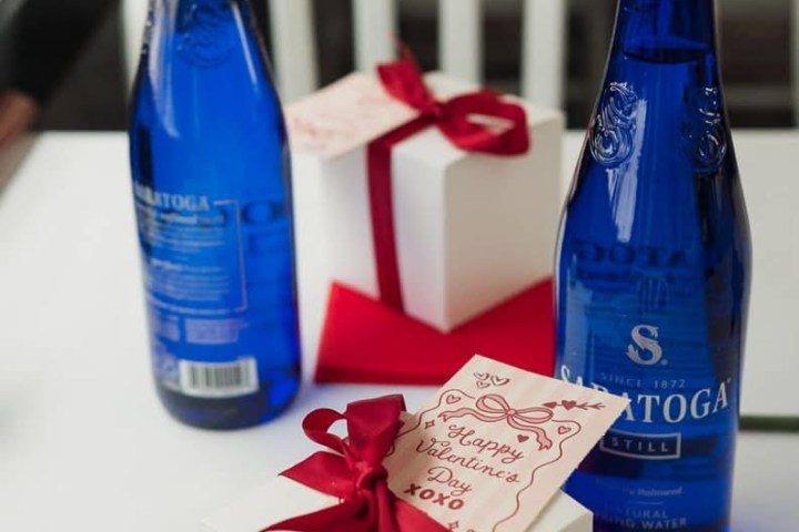Two blue bottles and two white gift boxes with red ribbons and cards on a table.