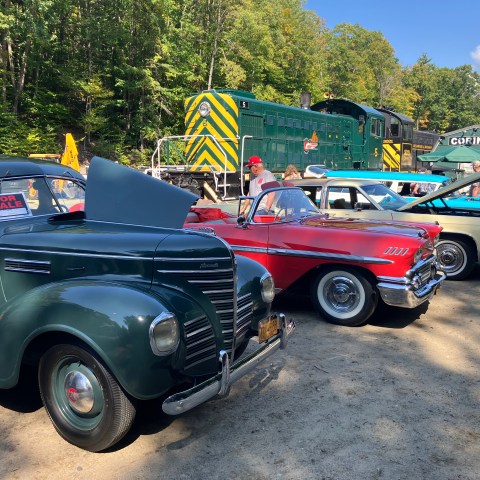 Classic cars parked outdoors with a vintage train in the background under a clear blue sky.