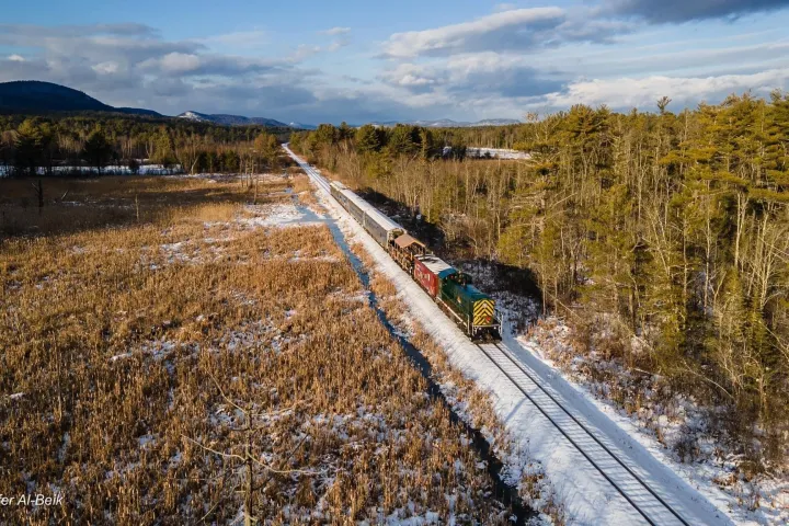 Aerial view of a train traveling through a snowy, forested landscape under a cloudy sky.