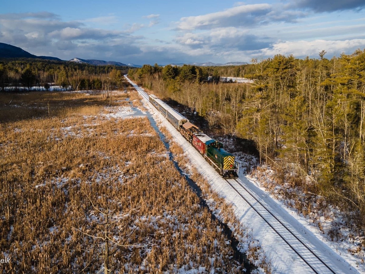 Aerial view of a train traveling through a snowy, forested landscape under a cloudy sky.