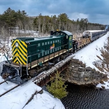 Train crossing a snowy bridge over a river with trees in the background.