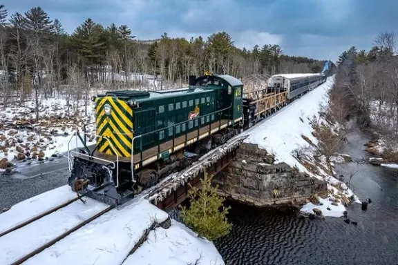 Train crossing a snowy bridge over a river with trees in the background.