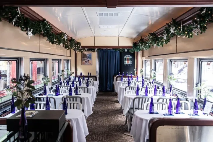 Decorated train dining car with white tablecloths, blue bottles, and garlands along windows.