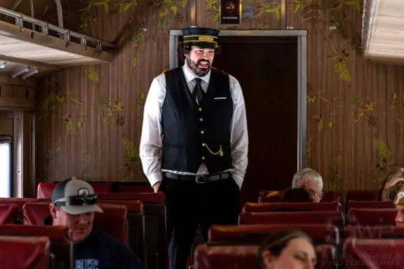 Train conductor in uniform stands inside a vintage train car with seated passengers.