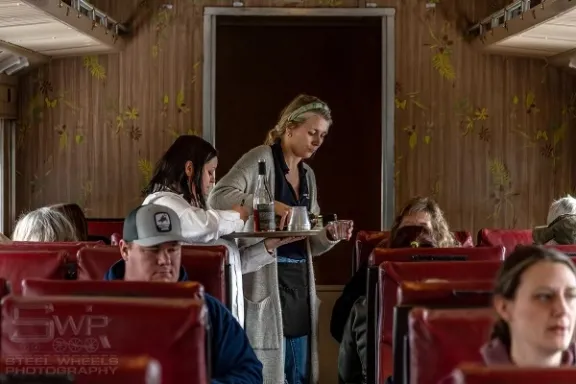 Train interior with passengers and server carrying drinks on a tray.
