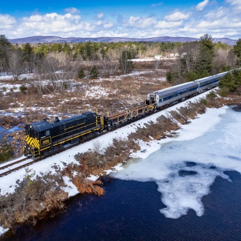 Train on tracks beside snowy landscape and partially frozen water, with trees and mountains in background.