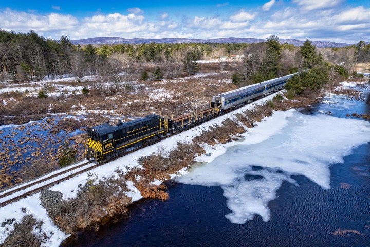 Train on tracks beside snowy landscape and partially frozen water, with trees and mountains in background.