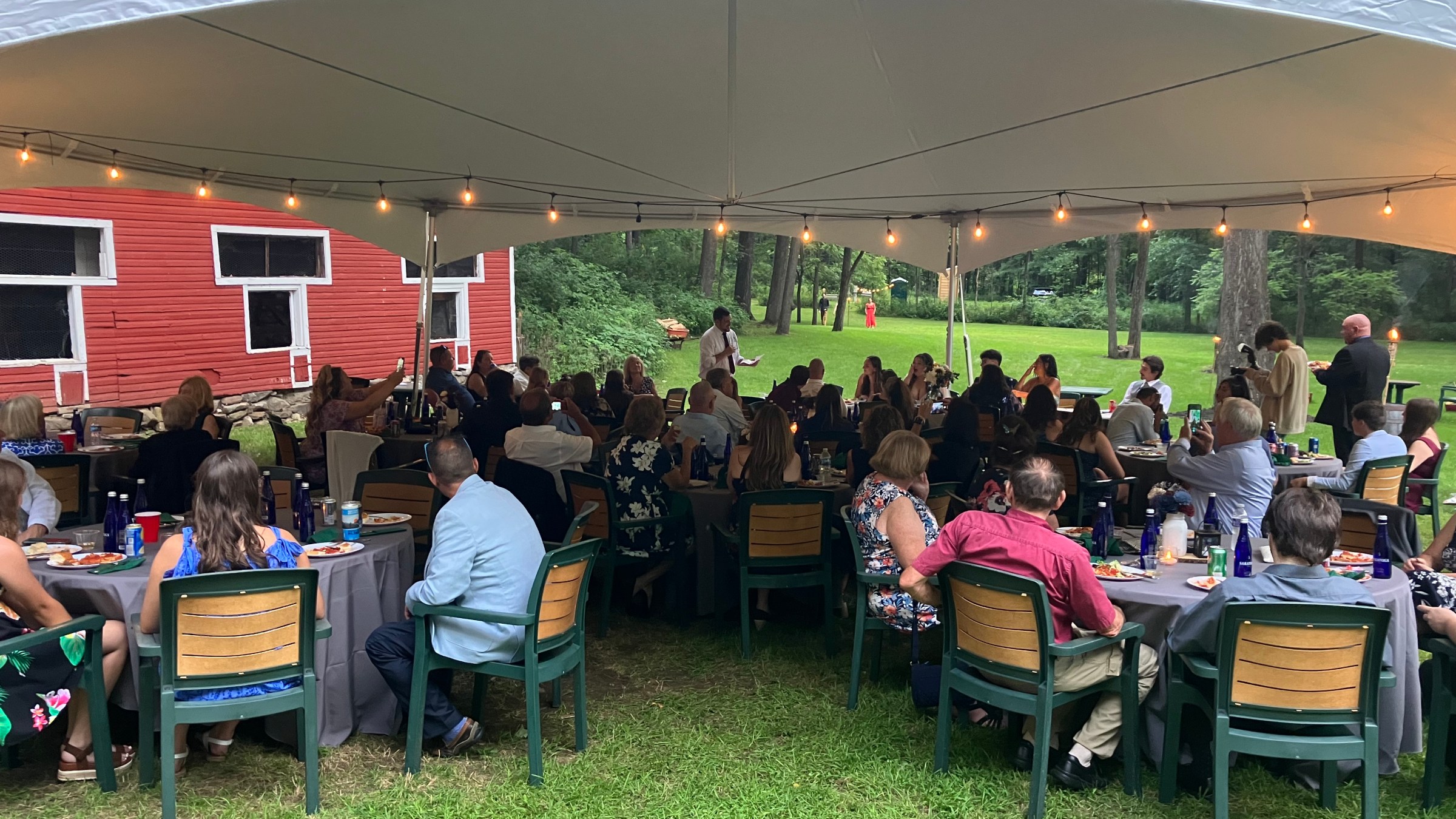 Outdoor gathering under a tent near a red barn with people seated at tables.