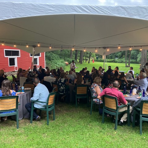 Outdoor gathering under a tent near a red barn with people seated at tables.