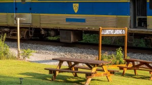 Train passes wooden tables near Lamothe Landing sign on grassy area.