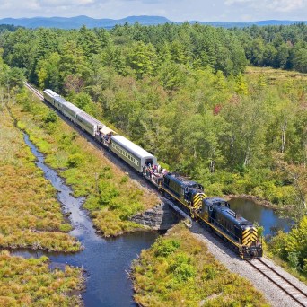 Aerial view of a train traveling through a green forested area with a river and blue sky.