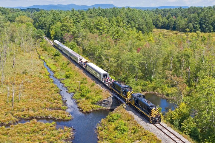 Aerial view of a train traveling through a green forested area with a river and blue sky.