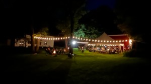 Night scene with string lights over a tent and people gathered around a campfire.