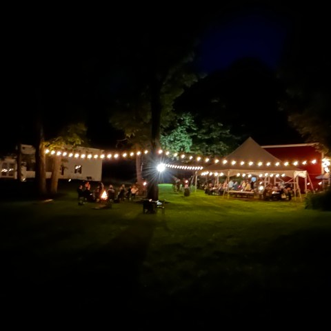 Night scene with string lights over a tent and people gathered around a campfire.