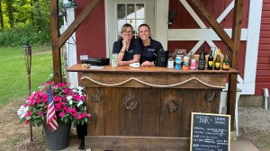 Two people behind an outdoor wooden bar with drinks and a menu board.