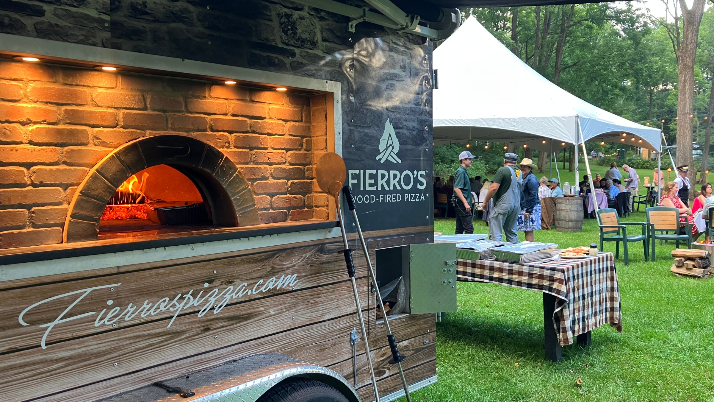 Pizza oven trailer at outdoor event with people under a white tent.