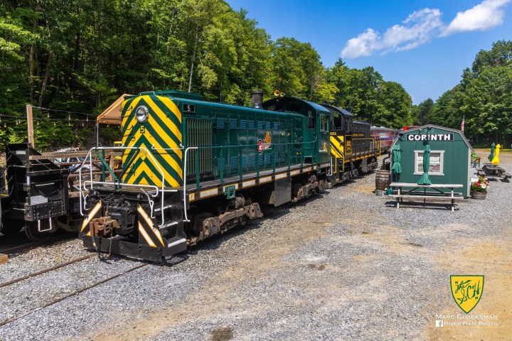 Green and yellow train at Corinth station surrounded by trees.
