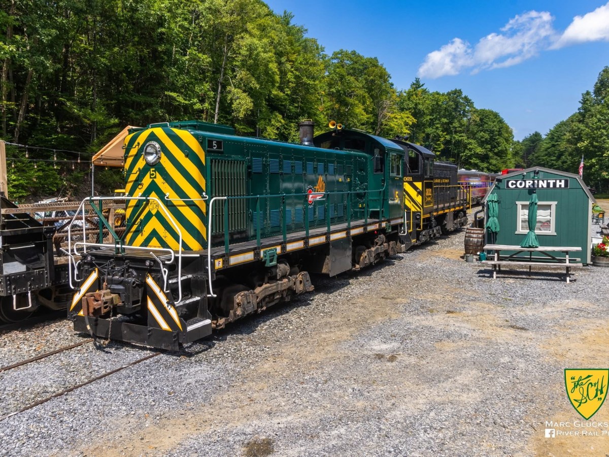 Green and yellow train at Corinth station surrounded by trees.