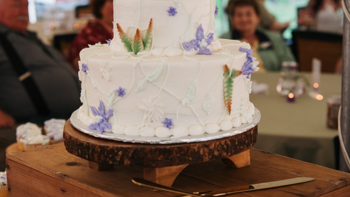 White wedding cake with floral decorations and a cake topper on a wooden stand.
