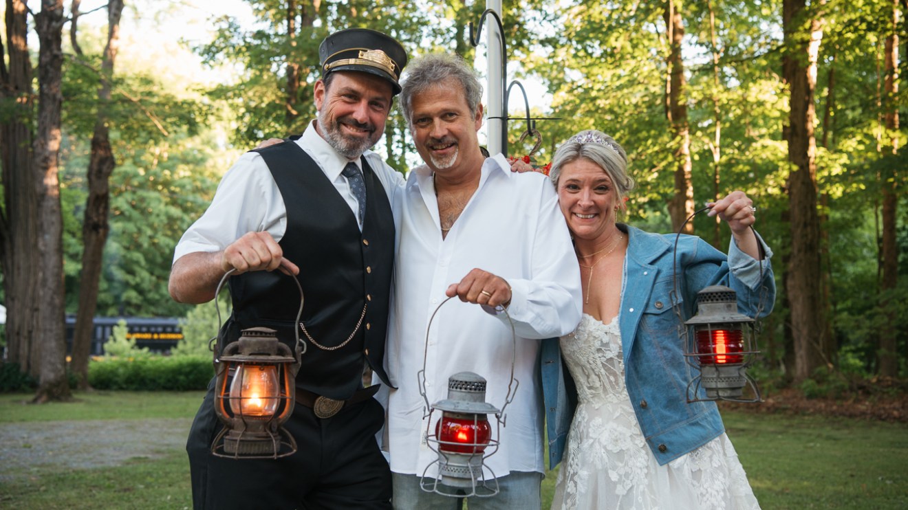 Three people outdoors holding lanterns, one in a conductor hat, one in a wedding dress.
