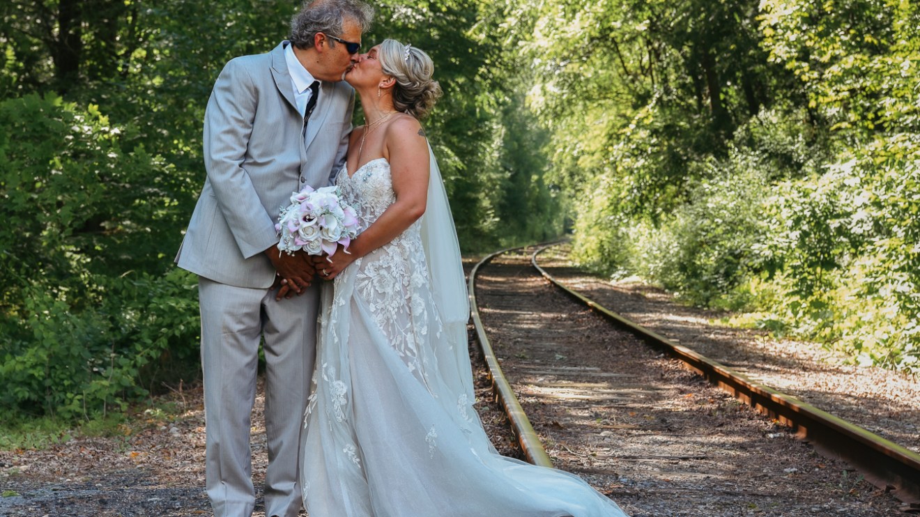 Bride and groom kissing on a railroad track surrounded by trees.