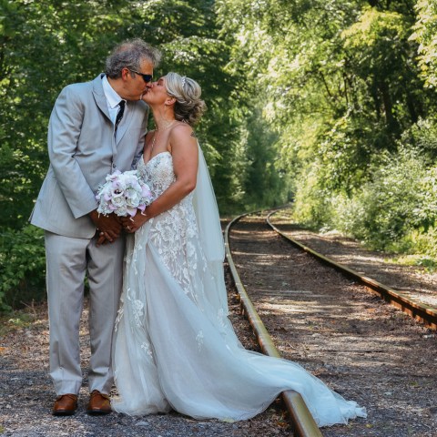 Bride and groom kissing on a railroad track surrounded by trees.